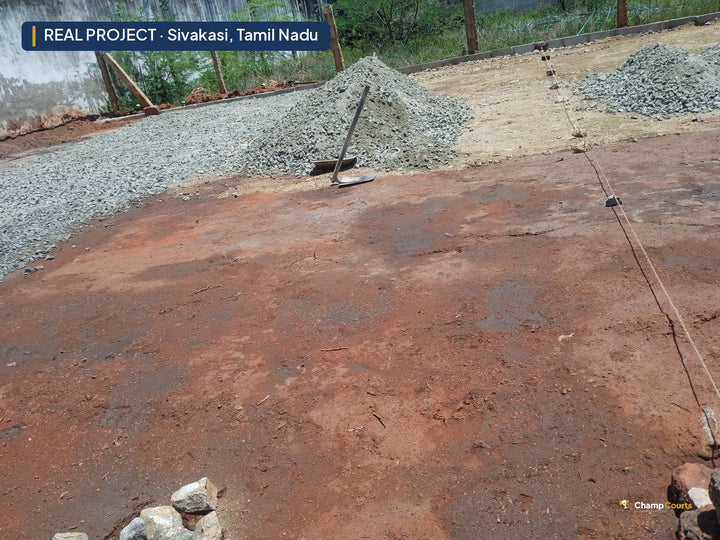 Bare red-earth plot in Sivakasi set out with string lines, stockpiles of grey aggregate along the boundary wall before b